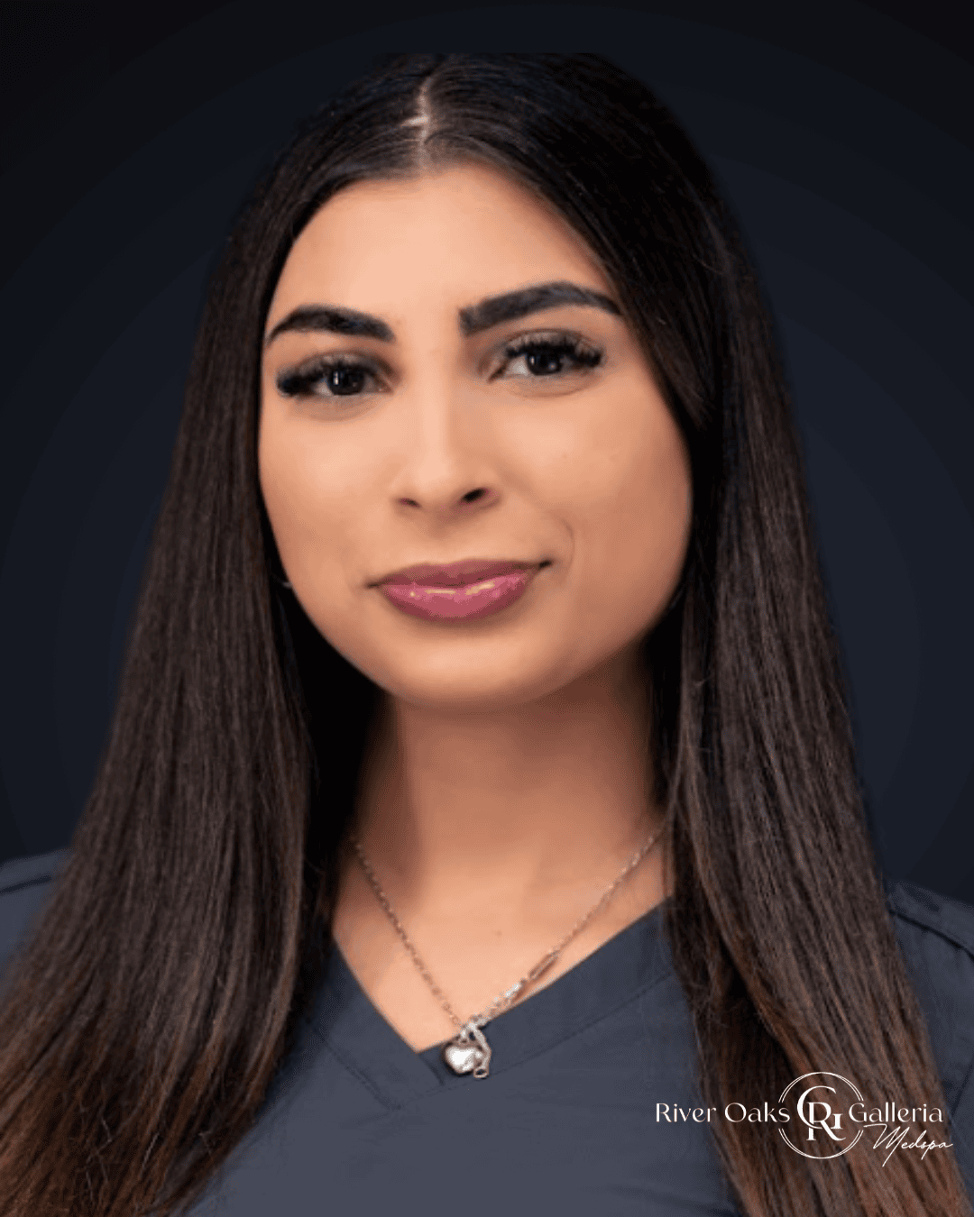 A woman with long dark hair wearing a navy blue top and a silver necklace with a heart-shaped pendan.