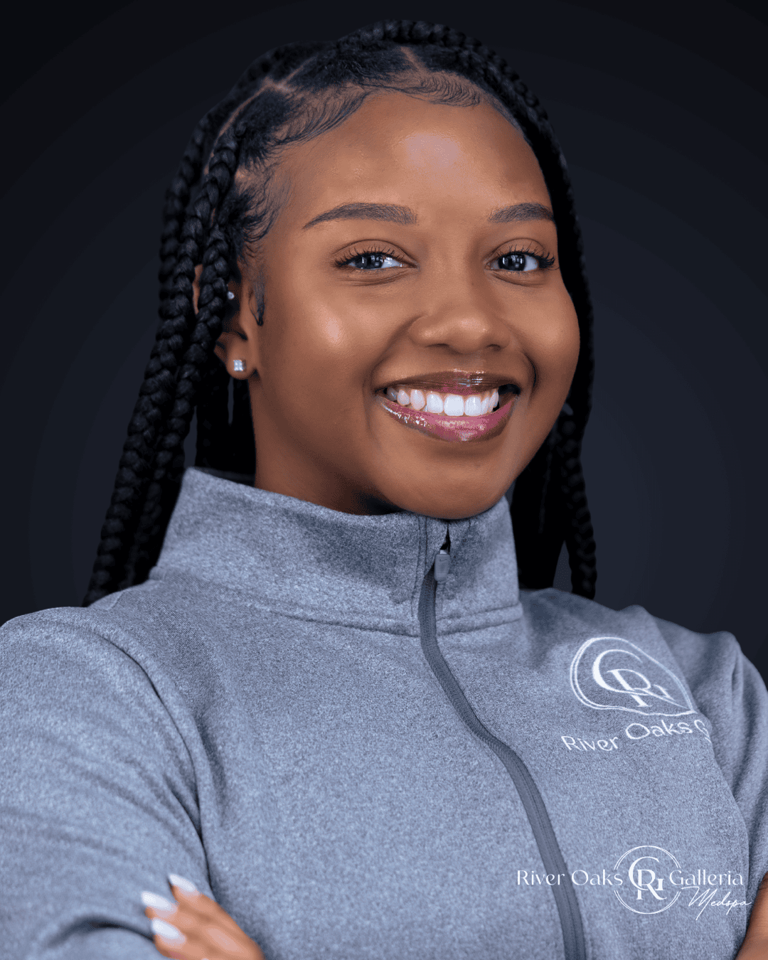 A smiling young woman with braided hair wearing a gray zip-up jacket with "River Oaks Galleria" logo.